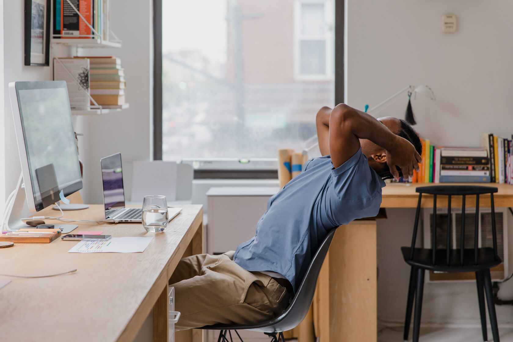 A man at a home office desk leaning back in his chair with both hands behind his neck